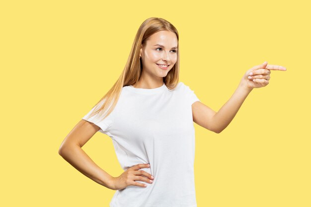 Caucasian young woman's half-length portrait on yellow studio background. Beautiful female model in white shirt. Concept of human emotions, facial expression. Pointing on side and smiling.