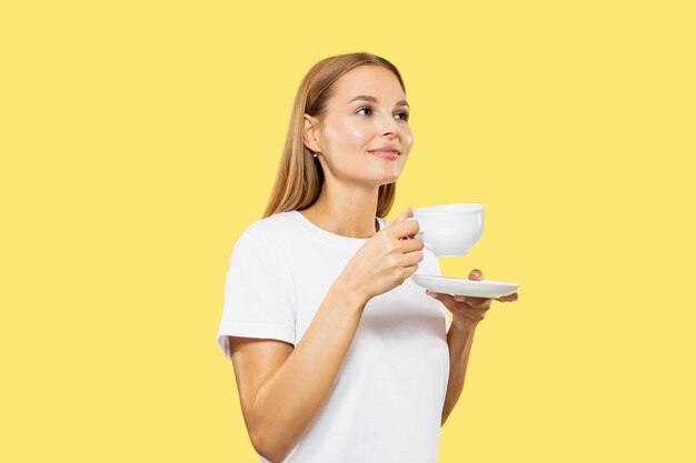 Caucasian young woman's half-length portrait on yellow studio background. Beautiful female model in white shirt. Concept of human emotions, facial expression, sales. Enjoing coffe or tea with cup.