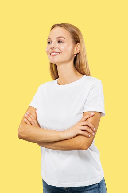 黄色のスタジオの背景に白人の若い女性の半身像。白いシャツの美しい女性モデル。人間の感情、表情、販売の概念。立っている手が交差し、自信を持っています。