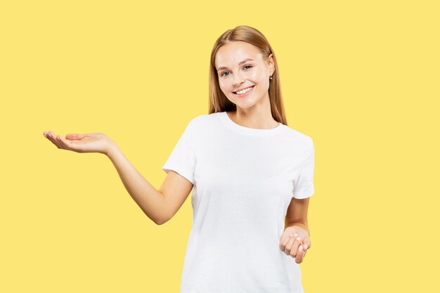 Caucasian young woman's half-length portrait on yellow studio background. Beautiful female model in white shirt. Concept of human emotions, facial expression. Showing and pointing something.