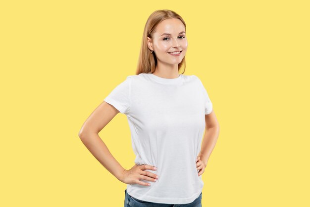노란색 스튜디오 배경에 백인 젊은 여자의 절반 길이 초상화. 흰 셔츠에 아름 다운 여성 모델입니다. 인간의 감정, 표정의 개념. 미소하고 엉덩이에 손으로 서.
