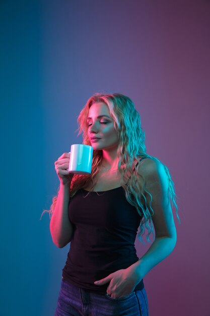 Caucasian young woman's portrait on gradient background in neon light. Beautiful female model with unusual look. Concept of human emotions, facial expression, sales, ad. Drinking coffee or tea.