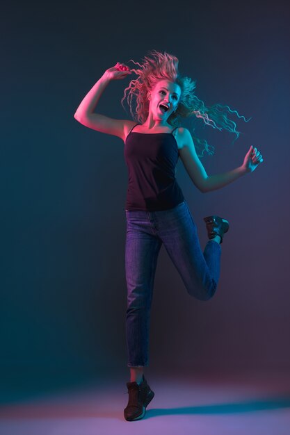 Caucasian young woman's portrait on gradient background in neon light. Beautiful female model with unusual look. Concept of human emotions, facial expression, sales, ad. Jumping, smiling.