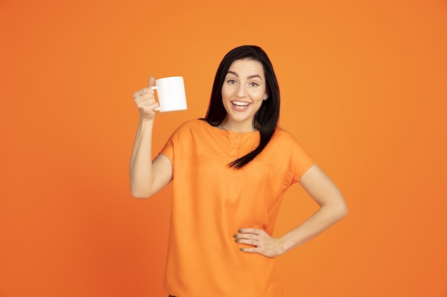 Caucasian young woman's portrait on orange studio background. Beautiful female brunette model in shirt. Concept of human emotions, facial expression, sales, ad. Copyspace. Drinking coffee or tea.