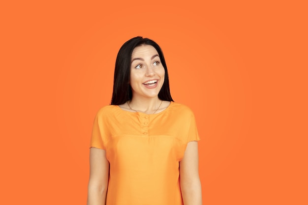 Портрет молодой женщины Кавказа на оранжевом фоне студии. Красивая женская модель брюнет в рубашке. Понятие человеческих эмоций, выражения лица, продаж, рекламы. Copyspace. Выглядит мечтательно, улыбается.