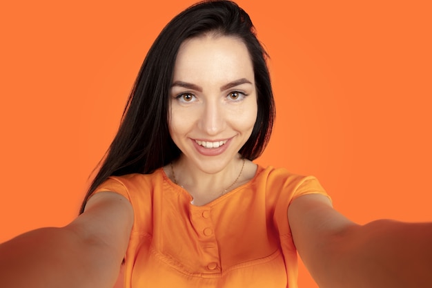 Портрет молодой женщины Кавказа на оранжевом фоне студии. Красивая женская модель брюнет в рубашке. Концепция человеческих эмоций, выражения лица, продаж, рекламы. Copyspace. Делаю селфи и улыбаюсь.