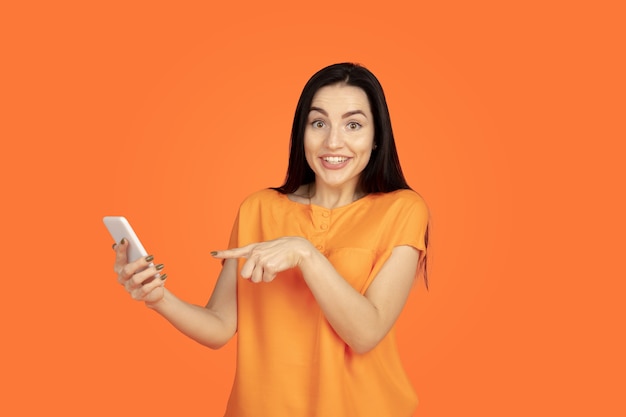 Caucasian young woman's portrait on orange studio background. beautiful female brunette model in shirt. concept of human emotions, facial expression, sales, ad. copyspace. making selfie, win in bet.