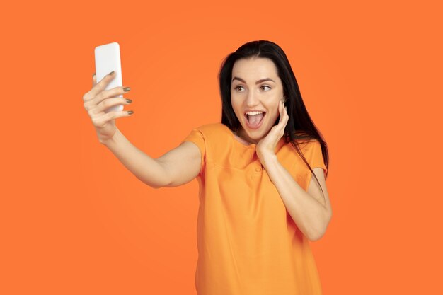 Caucasian young woman's portrait on orange studio background. Beautiful female brunette model in shirt. Concept of human emotions, facial expression, sales, ad. Copyspace. Making selfie, win in bet.