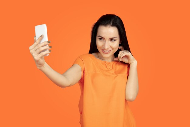 Портрет молодой женщины Кавказа на оранжевом фоне студии. Красивая женская модель брюнет в рубашке. Понятие человеческих эмоций, выражения лица, продаж, рекламы. Copyspace. Делаем селфи, выигрываем в ставке.