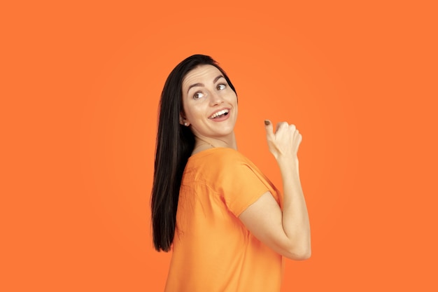 Caucasian young woman's portrait on orange studio background. Beautiful female brunette model in shirt. Concept of human emotions, facial expression, sales, ad. Copyspace. Pointing, showing, smiling.