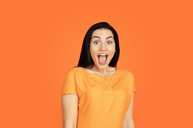 Caucasian young woman's portrait on orange studio background. Beautiful female brunette model in shirt. Concept of human emotions, facial expression, sales, ad. Copyspace. Shocked, astonished.