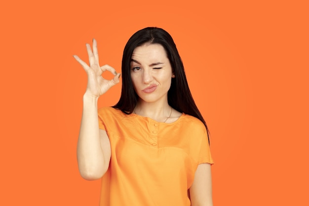 Портрет молодой женщины Кавказа на оранжевом фоне студии. Красивая женская модель брюнет в рубашке. Понятие человеческих эмоций, выражения лица, продаж, рекламы. Copyspace. Показывает знак ОК.