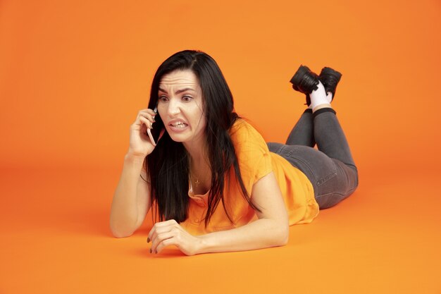 Caucasian young woman's portrait on orange studio background. Beautiful female brunette model in shirt. Concept of human emotions, facial expression, sales, ad. Copyspace. Talking on phone.