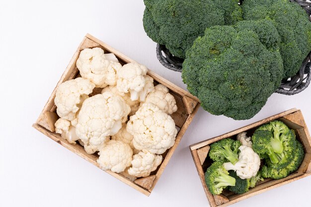 Cauliflower in box near the broccoli in basket top view on white surface