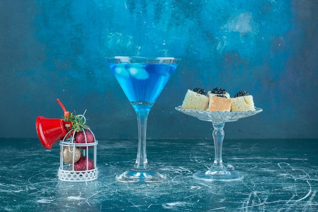 Caviar snacks on a glass pedestal next to a glass of cocktail and christmas ornaments on blue.