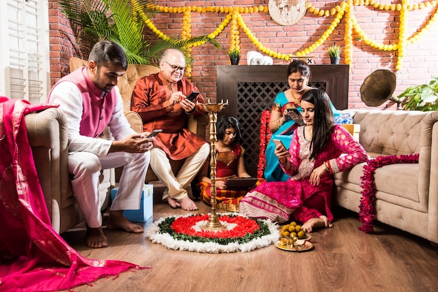 インドの携帯電話中毒-ディワリ祭でスマートフォンを使用する多世代のインドの家族、ディワリディヤ、花のランゴーリー、お菓子で飾られた家