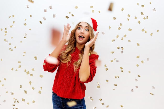 Celebrating girl in Santa masquerade hat having fun in confetti. New ear party mood. Cozy red pullover