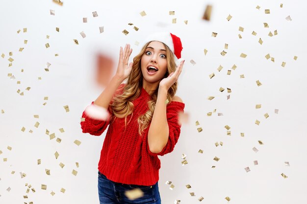 Celebrating girl in Santa masquerade hat having fun in confetti on white wall. New ear party mood. Cozy red pullover. True emotions. Surprise crazy emotions.