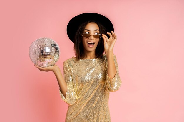 Celebrating woman in golden sequins dress posing with disco baloon over pink bacground in studio