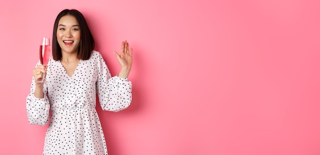 Free photo celebration beautiful asian woman drinking champagne and smiling standing in dress over pink backgro