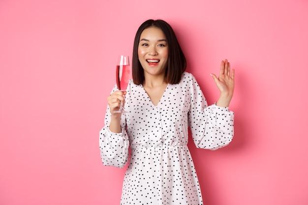 축하. 아름다운 아시아 여성이 샴페인을 마시고 웃고, 분홍색 배경 위에 드레스를 입고 서 있습니다.