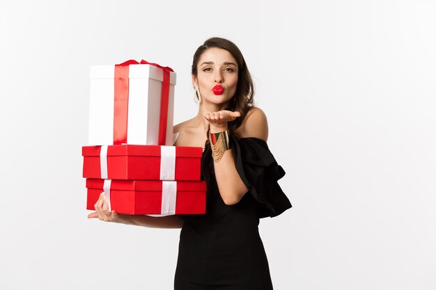 Концепция празднования и рождественских праздников. Красивая женщина в элегантном черном платье держит подарки, отправляя воздушный поцелуй в камеру, стоя на белом фоне.