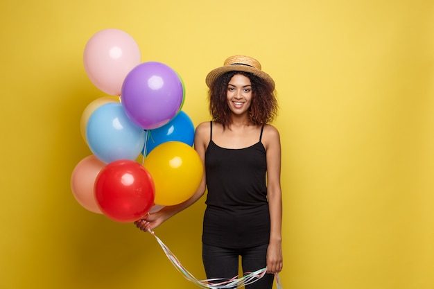 Celebration Concept - Close up Portrait happy young beautiful african woman in black t-shirt smiling with colorful party balloon. Yellow Pastel studio Background.