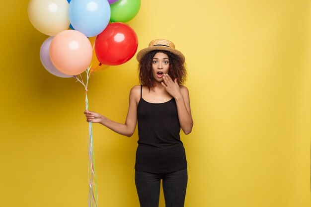 Celebration Concept - Close up Portrait happy young beautiful african woman in black t-shirt smiling with colorful party balloon. Yellow Pastel studio Background.