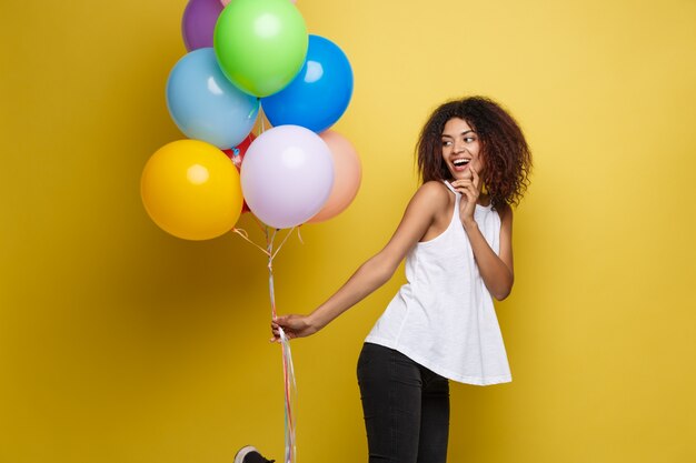 Празднование концепции - закрыть Портрет счастливый молодых красивых африканских женщина в черной футболке, улыбаясь с красочный участник воздушного шара. Желтая пастельная студия Фон.