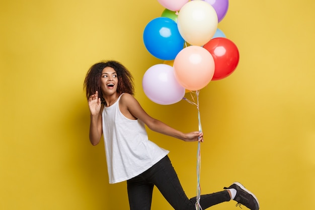 Free photo celebration concept - close up portrait happy young beautiful african woman in black t-shirt smiling with colorful party balloon. yellow pastel studio background.