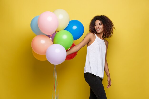 Celebration Concept - Close up Portrait happy young beautiful african woman in black t-shirt smiling with colorful party balloon. Yellow Pastel studio Background.