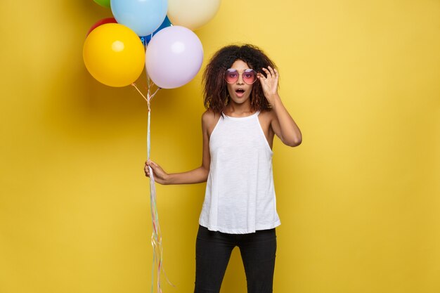 Celebration Concept - Close up Portrait happy young beautiful african woman in black t-shirt smiling with colorful party balloon. Yellow Pastel studio Background.