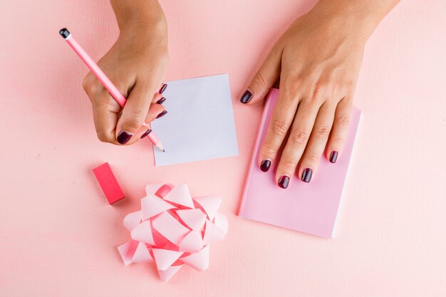 Celebration concept with bow, mini notebook, eraser on pink table flat lay. woman writing on sticky note.