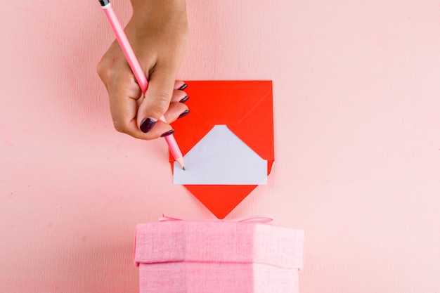 Celebration concept with gift box on pink table flat lay. woman signing greeting card.