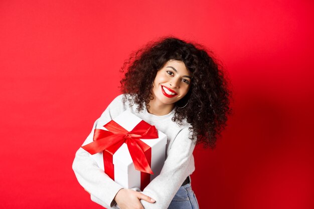 Celebration and holidays concept. Happy woman holding birthday gift and smiling at camera, standing in casual clothes, red background