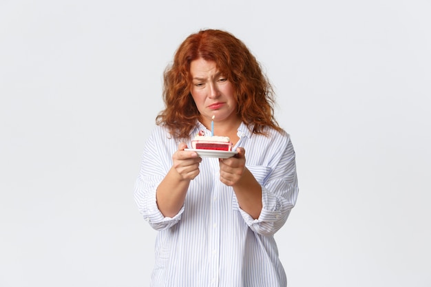 Celebration, holidays and emotions concept. Uneasy and sad redhead middle-aged woman hate her birthday, looking upset at b-day cake with lit candle, feeling mid-life crisis, white wall.