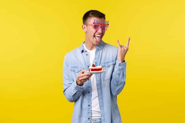 Celebration, holidays and people emotions concept. Carefree happy blond guy celebrating birthday, enjoying party, show rock-on sign and holding b-day cake, yellow background,