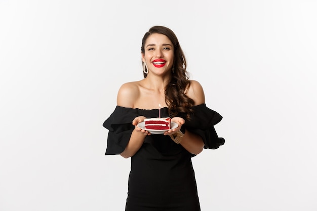 Celebration and party concept. Happy gorgeous woman having birthday, holding b-day cake and smiling, making wish, standing in black dress with makeup.