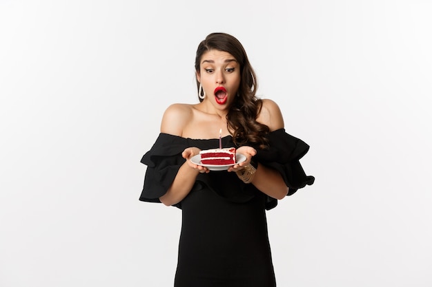 Celebration and party concept. Happy woman blowing candle on birthday cake, making wish, standing in black dress and makeup against white background.