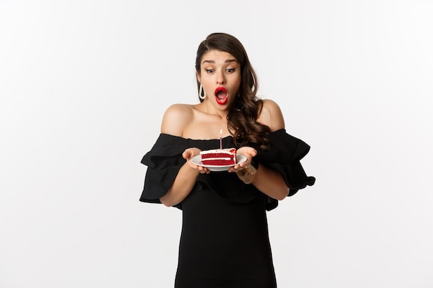 Celebration and party concept. Happy woman blowing candle on birthday cake, making wish, standing in black dress and makeup against white background