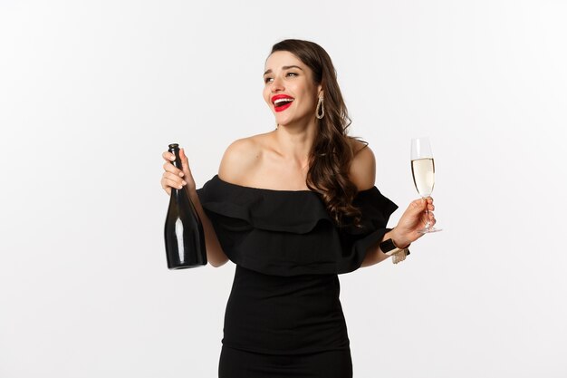 お祝いとパーティーのコンセプト。新年の休暇を楽しんで、シャンパンのボトルとグラスを保持している魅力的なドレスを着たスタイリッシュなブルネットの女性。
