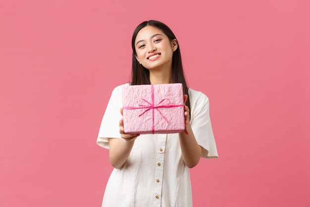 축하, 파티 휴일 및 재미있는 개념입니다. 친절한 아름다운 아시아 여자 친구가 웃고, 생일을 축하하고 선물을 주고 분홍색 배경에 서 있습니다.