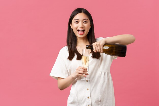 Празднование, партийные праздники и забавная концепция. Счастливая беззаботная азиатская женщина в отпуске, наливает шампанское в бокал и смеется, наслаждаясь выходными или отдыхом, стоя на розовом фоне.