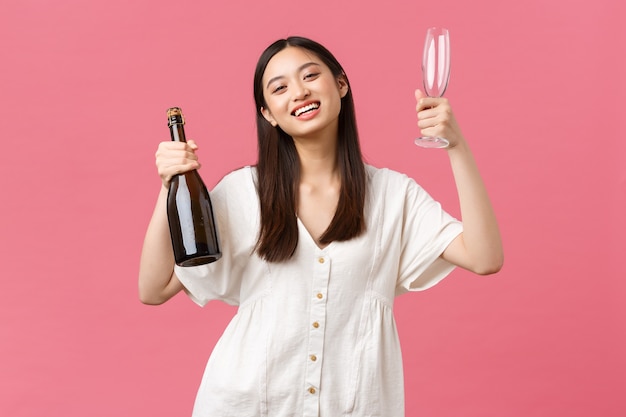 Celebrazione, feste di festa e concetto di divertimento. gioiosa ragazza asiatica felice pronta a godersi il giorno libero con le amiche, portare champagne e bicchieri, macchina fotografica sorridente, in piedi sfondo rosa ottimista.