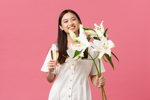 Celebration, party holidays and fun concept. Silly happy birthday girl in white dress, smiling broadly as receive beautiful bouquet of lilies, holding glass of champagne, standing pink background
