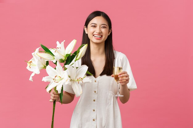 Celebration, party holidays and fun concept. Silly happy birthday girl in white dress, smiling broadly as receive beautiful bouquet of lilies, holding glass of champagne, standing pink background.