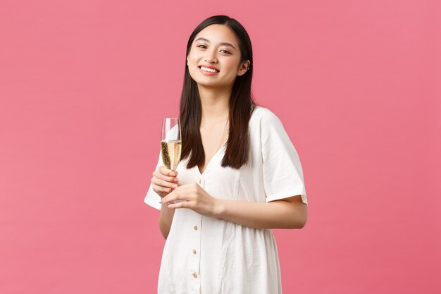 Празднование, партийные праздники и забавная концепция. Улыбаясь с днем рождения девушка в белом платье, наслаждаясь празднованием с друзьями, держа бокал шампанского на розовом фоне. Копировать пространство