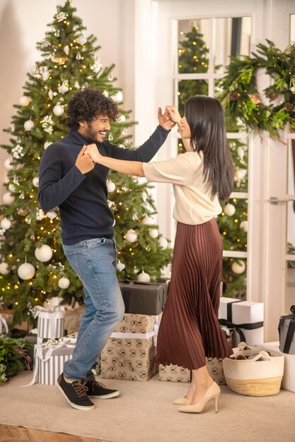 Celebration. Young bearded smiling indian man and pretty asian woman in high heels looking at each other dancing touching hand near christmas tree and gifts