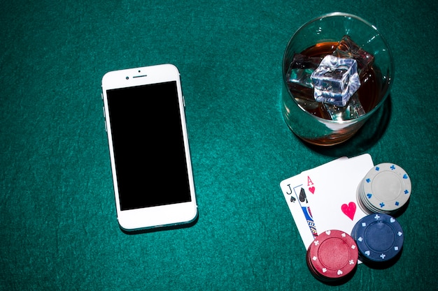 Бесплатное фото Мобильный телефон и стекло виски с гнездом лопаты и сердечными тузами на зеленом покерном столе