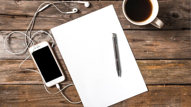 Cellphone connected with earphones; pen; paper and coffee cup on wooden background